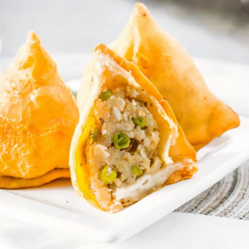 Golden-brown samosas on a white plate, one cut open to reveal a savory potato and pea filling.