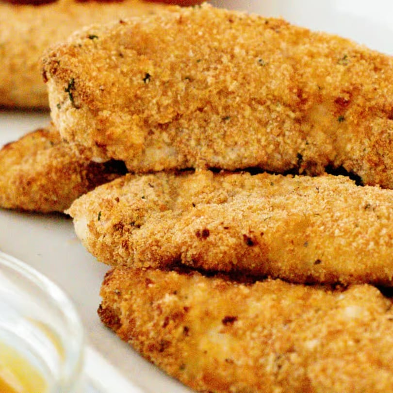 Close-up of golden-brown, breaded chicken tenders or fish sticks, stacked on a white plate.