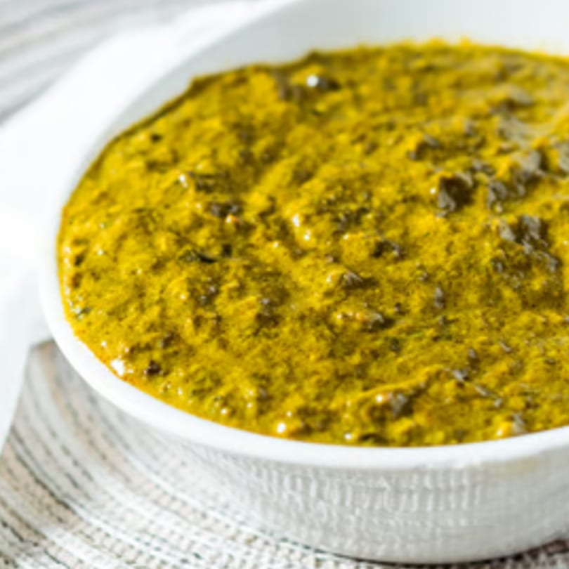 A white bowl filled with vibrant green Indian curry, possibly saag, on a patterned placemat.