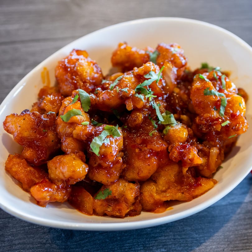 A white bowl filled with glistening orange-red Indian Palace Roseville Gobi Manchurian, garnished with fresh cilantro.